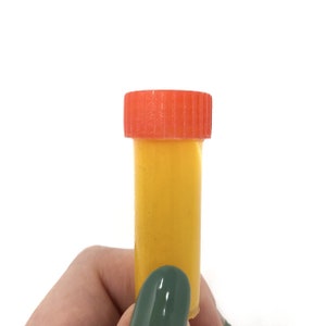 May include: A small, yellow plastic container with a bright orange lid. The container is approximately 5 cm tall and 2.5 cm in diameter.