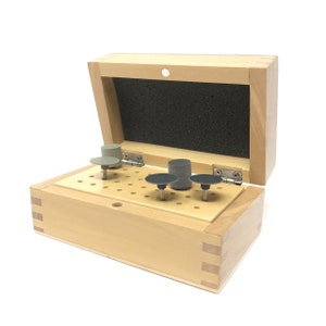 May include: A wooden box with a hinged lid, open to reveal a set of small, round sanding tools. The tools are arranged in a grid of holes in the box's interior. The tools are various shades of gray and black.