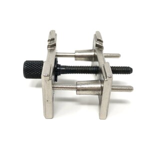 Puede incluir: Una herramienta de sujeci&oacute;n de correa de reloj de plata con una perilla negra. La herramienta tiene dos brazos ajustables con pasadores que se pueden usar para sujetar una correa de reloj en su lugar mientras se trabaja.