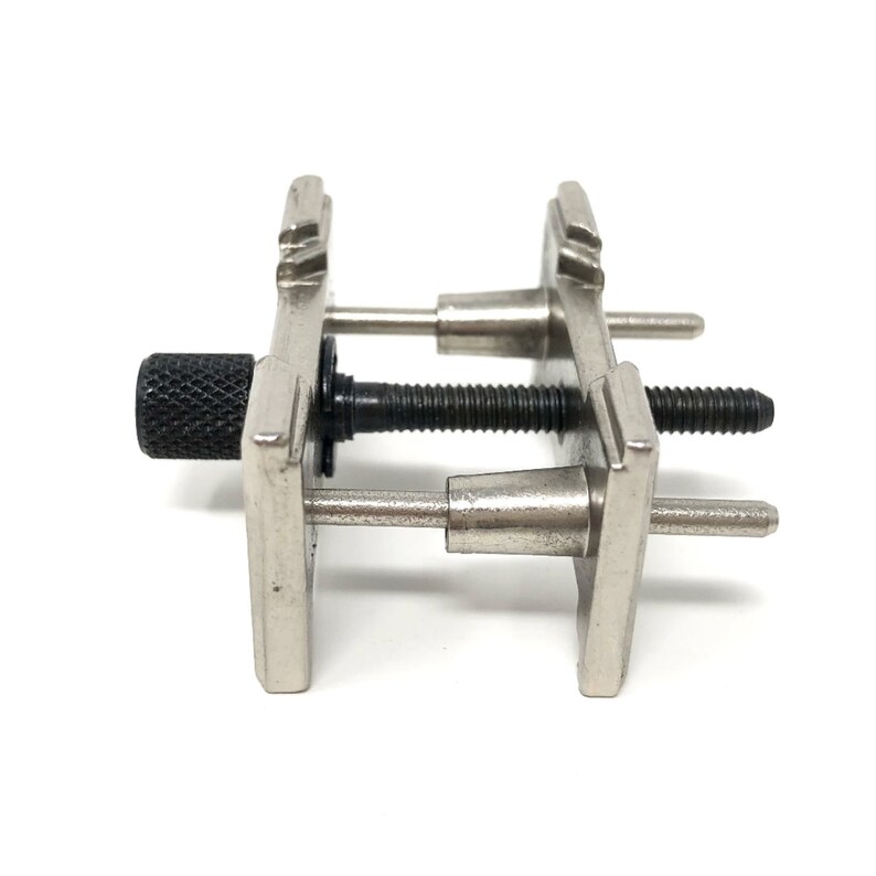 Puede incluir: Una herramienta de sujeci&oacute;n de correa de reloj de plata con una perilla negra. La herramienta tiene dos brazos ajustables con pasadores que se pueden usar para sujetar una correa de reloj en su lugar mientras se trabaja.