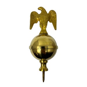 May include: A gold-coloured metal flag pole topper featuring an eagle perched atop a sphere. The eagle has outstretched wings and is detailed with feathers. The sphere has a horizontal band around the middle. A screw extends from the bottom.