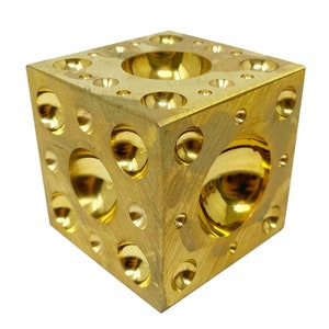 May include: A gold metal cube with a textured surface. The cube has multiple round indentations of varying sizes.