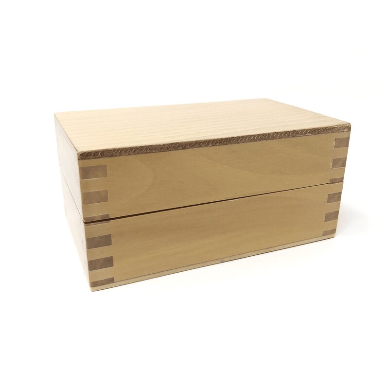 May include: A wooden box with a light brown finish. The box has a hinged lid and is made of two pieces of wood that are joined together with a dovetail joint.