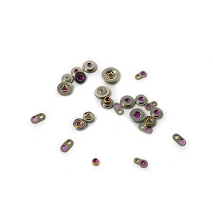 May include: Assortment of small, silver-toned metal parts with pink gemstones. Some parts are round, some are oval, and some are shaped like triangles. The parts are scattered on a white surface.