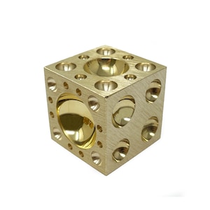 May include: A gold metal cube with multiple rounded indentations on each side. The cube has a large, rounded indentation on the top.