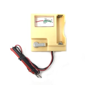May include: A cream-coloured cell tester with a battery compartment and a gauge displaying 1.5V and 3V readings. It has a metal switch and is connected to red and black wires with metal probes. The text "CELL TESTER" is visible.