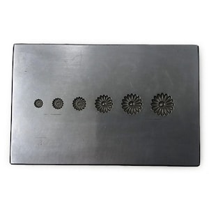 May include: A rectangular, dark grey metal stamping block featuring six daisy-shaped impressions of varying sizes. The impressions are arranged in a row, increasing in size from left to right. The block is likely used for metalworking or jewellery making.