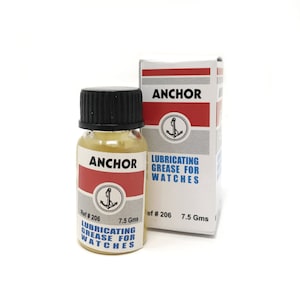 Puede incluir: Una peque&ntilde;a botella de vidrio de grasa lubricante Anchor para relojes, con tapa negra y etiqueta blanca y roja. La etiqueta incluye el nombre de la marca, un logotipo de ancla y el texto "Grasa lubricante para relojes". Una caja a juego est&aacute; detr&aacute;s de la botella.