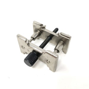 Puede incluir: Una herramienta de sujeci&oacute;n de correa de reloj de plata con una perilla negra. La herramienta se utiliza para sujetar una correa de reloj en su lugar mientras se trabaja en ella.