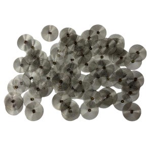May include: A cluster of small, silver-coloured, circular metal springs with a spiral design. Each spring has a central, darker-coloured hub. The springs are scattered across a white background, creating a textured, industrial aesthetic.
