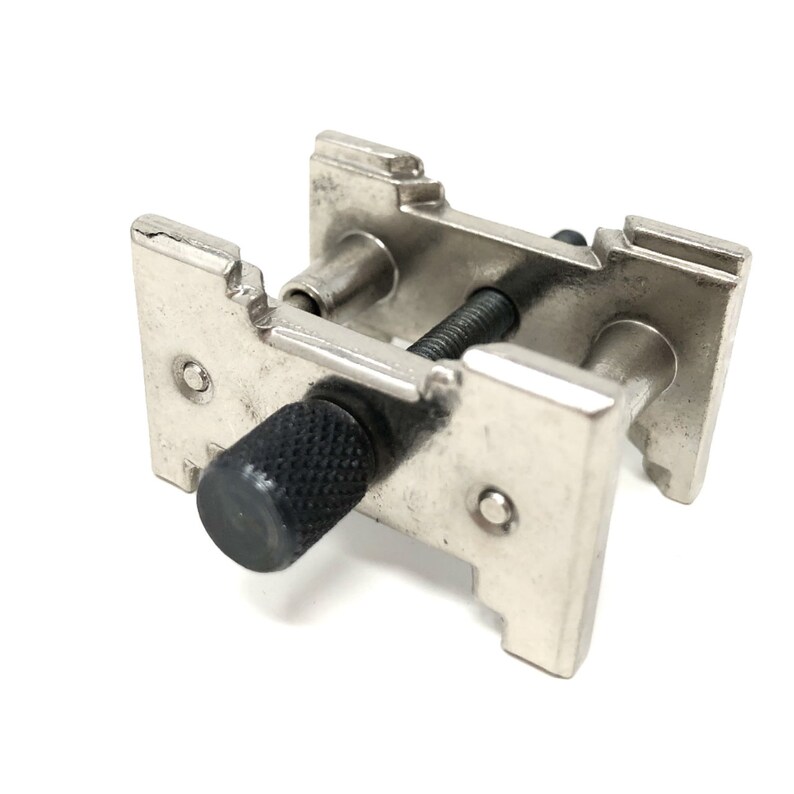 Puede incluir: Una herramienta para correas de reloj de metal plateado con una perilla negra. La herramienta tiene dos brazos ajustables con dientes para sujetar la correa del reloj en su lugar mientras se ajusta.