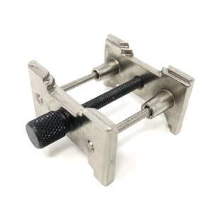 Puede incluir: Una herramienta de prensa para correas de reloj de plata con una perilla negra. La herramienta se utiliza para quitar e instalar correas de reloj.