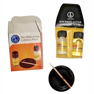 Puede incluir: Un juego de pasta luminiscente no radiactiva en una caja pequeña con dos botellas transparentes y un palillo de dientes. La caja tiene un fondo amarillo con el texto "Pasta luminiscente no radiactiva" y un pequeño logotipo de ancla. Las botellas están etiquetadas con "Anchor" y "CHOF US POWD DIOACTIVE".