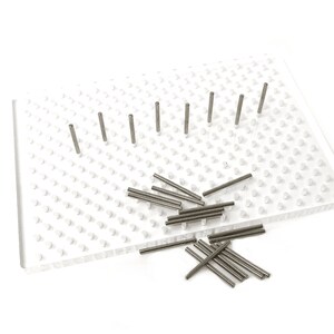 May include: A clear plastic pegboard with numerous small pegs and several metal pegs inserted. A pile of loose metal pegs is scattered on the board. The pegs are cylindrical and silver in colour. The board is rectangular.