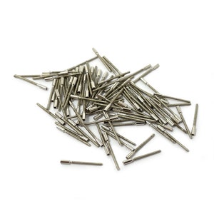 May include: A pile of silver-coloured metal pins with rounded heads. The pins are scattered on a white surface.