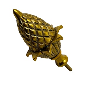 May include: A gold-coloured, pineapple-shaped decorative finial. The finial has a textured surface with a screw at the base. The pineapple design is detailed with leaves and a pointed top. The overall design is ornate and decorative.