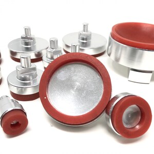 May include: A set of eight silver-colored metal bases with red rubber tops. The bases have a small knob on top and are designed to be used as a base for crafting projects.