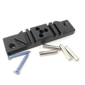 May include: Black metal jig with multiple slots and holes for woodworking. The jig is accompanied by five silver metal dowel pins and two blue screws.