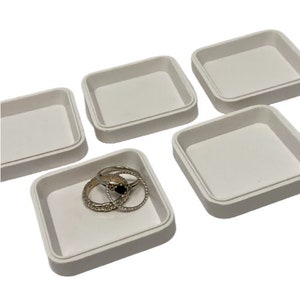 May include: Five square, off-white jewelry trays. One tray contains several silver rings with intricate designs and a black gemstone. The other four trays are empty. The trays are made of a smooth, matte material.