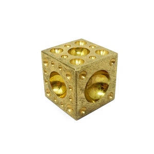 May include: A brass cube featuring a pattern of circular holes and spherical indentations. The cube has a polished, reflective surface. The holes are of varying sizes. The design is geometric and modern.