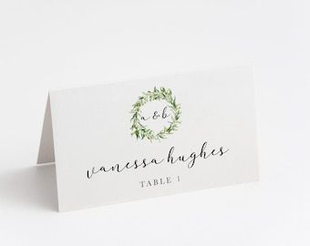 May include: White wedding place card with a green leafy wreath with the initials "A & B" in the center. The text "vanessa hughes" and "TABLE 1" are printed below the wreath.