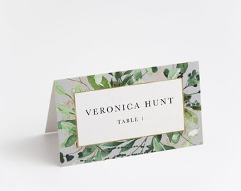 May include: A white place card with a gold frame and green watercolor floral border. The text "VERONICA HUNT TABLE 1" is printed in black.
