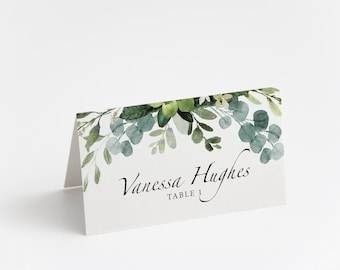 May include: A white place card with a green watercolor floral design. The text "Vanessa Hughes Table 1" is printed in black.