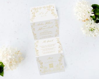 May include: A folded wedding invitation with gold floral accents and elegant script. The invitation is open, revealing details about the event. White flowers and greenery are arranged around the invitation on a white surface.