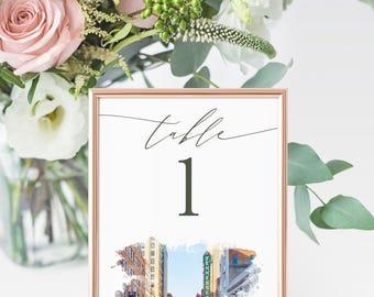 May include: A framed table number one with a watercolor street scene. The frame is rose gold. The words "table" and the number "1" are in a dark green script. A bouquet of pink and white flowers is in the background.