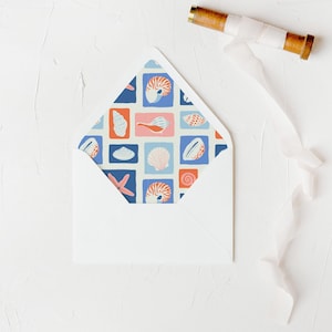 May include: A white envelope with a colorful interior pattern featuring seashells, starfish, and other marine life in a grid design. A wooden spool with white ribbon is in the upper right corner.