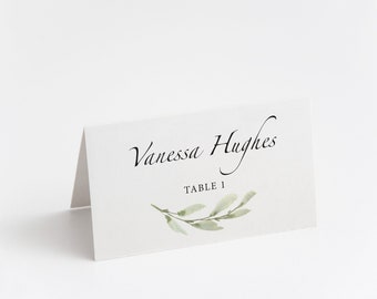 May include: White place card with black calligraphy that reads "Vanessa Hughes" and "Table 1". A green watercolor branch with leaves is at the bottom of the card.