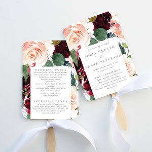 May include: Wedding ceremony program fans with a floral design featuring burgundy, pink, and green watercolor roses. The fans are tied with white ribbon and have a wooden handle. The text on the fans reads "Welcome to the wedding of Julia Hunter & Frank Peterson Saturday, the sixteenth of July  Lonesomeville, Ohio THE CEREMONY  Prelude - Seating of the families  The Ceremony - Exchange of vows  Exchange of rings - Reading  Presentation of newly married couple  Reception"