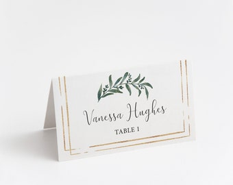 May include: White place card with gold trim and a green leafy branch design. The text on the card reads "Vanessa Hughes, Table 1."