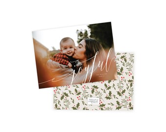 May include: Holiday card featuring a family photo with the word "joyful" in elegant script. The card's design includes a photo of a mother kissing a baby. The envelope has a pattern of green holly leaves and red berries.