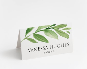 May include: A white place card with a green watercolor leaf design. The text "Vanessa Hughes Table 1" is printed in black.