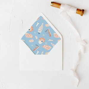 May include: A white envelope with a blue patterned interior featuring illustrations of seashells and other marine life. A wooden spool with white ribbon is in the upper right corner. The envelope is on a textured white surface.