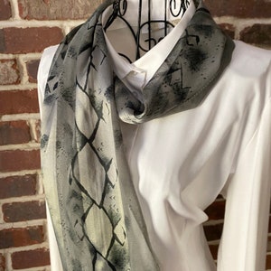 May include: A grey and black patterned silk scarf with a geometric design. The scarf is draped around a white shirt.