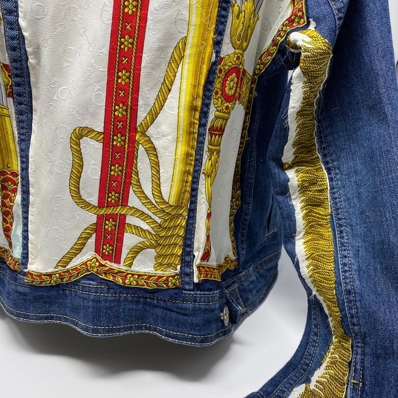 Peut inclure: Une veste en jean bleue avec un tissu &agrave; motifs blanc et or cousu sur le devant et les manches. Le tissu pr&eacute;sente un motif de corde et une bande rouge et blanche.