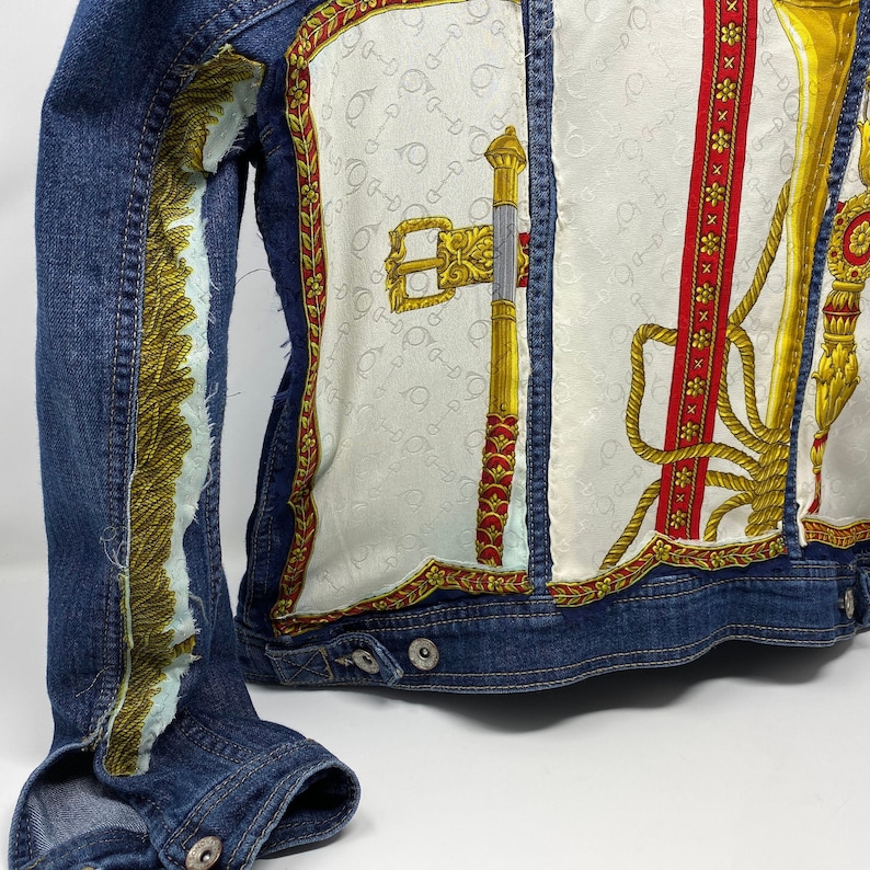 Peut inclure: Une veste en jean bleue avec un panneau de tissu &agrave; motifs blancs et dor&eacute;s sur le devant. Le panneau de tissu pr&eacute;sente un motif r&eacute;p&eacute;t&eacute; d'une cl&eacute; et d'un motif de corde.