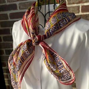 Peut inclure: Un foulard coloré avec un motif géométrique dans les tons de rouge, jaune, vert et violet. Le foulard est noué et drapé sur une chemise blanche, mettant en valeur son design complexe et ses couleurs vives. Le motif comprend des rayures et des cercles.