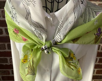 May include: A silk scarf with a white background and a green border, featuring floral and botanical designs. The scarf is knotted and worn over a white collared shirt. The scarf's patterns include black line drawings of trees and flowers.