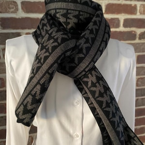 Op de afbeelding: Een zwarte en grijze sjaal met een herhalend patroon van in elkaar grijpende letters. De sjaal is over een wit overhemd gedrapeerd. Het ontwerp van de sjaal heeft een rand van grijze strepen. De achtergrond is een bakstenen muur.