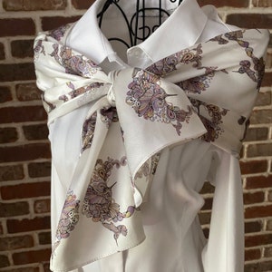Op de afbeelding: Een witte zijden sjaal met een kolibrie- en paisley-patroon is om een wit overhemd met kraag geknoopt. Het patroon van de sjaal herhaalt zich in paars, goud en groen. Het overhemd is dichtgeknoopt en de sjaal is aan de voorkant geknoopt.