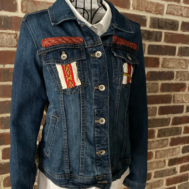 Peut inclure: Une veste en jean bleu fonc&eacute; avec des bordures rouges et dor&eacute;es et des empi&egrave;cements en tissu d&eacute;coratifs sur la poitrine.