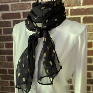 May include: A sheer black scarf featuring a repeating pattern of small, metallic gold dots. The lightweight fabric is tied in a knot at the centre, displaying a translucent texture and a consistent gold-flecked design across the entire surface.