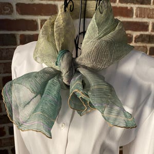 May include: A sheer, patterned scarf in shades of green, gold, and lavender is knotted. The scarf has a rippled texture and is draped over a white shirt. The edges of the scarf are trimmed with a gold border. The scarf is displayed on a black metal stand.