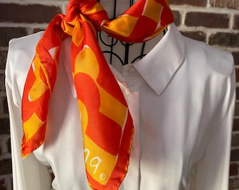 May include: A vibrant orange and yellow patterned scarf is draped over a white collared shirt. The scarf features a bold geometric design with the word "na" visible. The scarf is knotted at the neck, creating a stylish accessory.