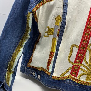 Peut inclure: Une veste en jean bleue avec un patch en tissu &agrave; motifs blancs, rouges et dor&eacute;s au dos. Le patch en tissu pr&eacute;sente un motif de corde, une &eacute;p&eacute;e et une bordure florale.