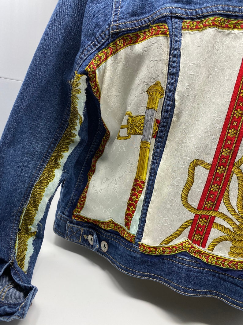 Peut inclure: Une veste en jean bleue avec un patch en tissu &agrave; motifs blancs, rouges et dor&eacute;s au dos. Le patch en tissu pr&eacute;sente un motif de corde, une &eacute;p&eacute;e et une bordure florale.