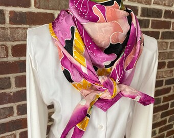 May include: A pink, yellow, and black silk scarf with a floral pattern. The scarf is tied around the neck of a white shirt.